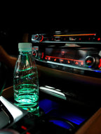 Coaster glowing green under a water bottle in a car cup holder beside a red-lit centre console at night